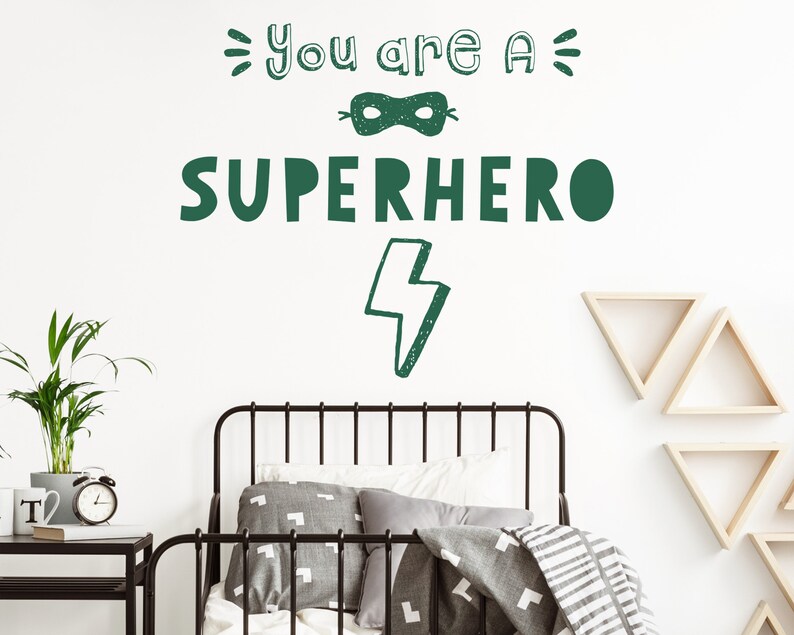 May include: Green and white wall decal with the text "You are a Superhero" and a lightning bolt. The decal is on a white wall above a black metal bed frame with grey bedding.