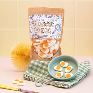 Op de afbeelding: Een bruin papieren zakje gevuld met gummisnoepjes in de vorm van gebakken eieren. Het label bevat de tekst "YOU'RE SUCH A GOOD EGG" omringd door illustraties van eieren in pannen. Een kleine groene pan bevat zes eivormige snoepjes op een geruite doek.