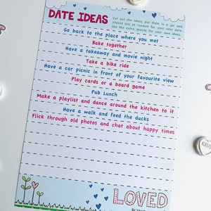 May include: A white card with blue and pink hearts and the text "Date Ideas" printed on it. The card has a list of date ideas, such as "Go back to the place where you met" and "Have a walk and feed the ducks". The card also has the text "Loved Beyond Words" printed on it.