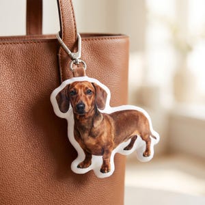 May include: A brown dachshund dog keyring with a silver clasp attached to a brown leather bag. The dog keyring is a plush, soft material with a detailed image of the dog.
