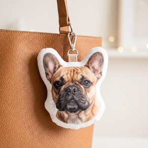 May include: A French Bulldog-shaped plush keychain with a realistic image of the dog's face. The keychain is attached to a brown leather bag with a silver clasp.