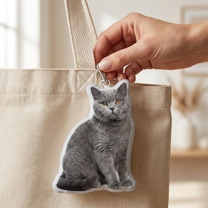Op de afbeelding: Een grijze katten sleutelhanger met een zilveren sluiting wordt voor een beige draagtas gehouden. De kat is een British Shorthair ras, met gele ogen en een gedetailleerd vachtpatroon. De sleutelhanger heeft de vorm van een kat.