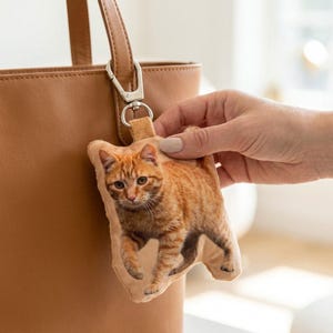 May include: A soft keychain with an orange tabby cat design. The keychain is attached to a brown leather tote bag with a silver clasp. The cat has a detailed fur pattern and is in a walking pose.
