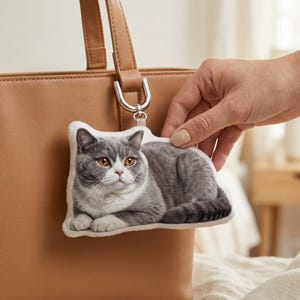 Op de afbeelding: Een pluche sleutelhanger met een grijze en witte British Shorthair kat met oranje ogen. De katvormige bedel is bevestigd aan een zilverkleurige metalen ring en wordt vastgehouden in de buurt van een bruine leren handtas.
