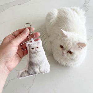 Op de afbeelding: Een witte katten sleutelhanger met een zilveren sluiting wordt vastgehouden naast een pluizige witte kat. De sleutelhanger heeft een foto van een kat met lichtbruine ogen en een grijs en witte vacht. De achtergrond is een wit marmeren oppervlak.