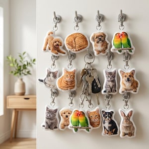 Op de afbeelding: Een verzameling sleutelhangers met verschillende dieren, waaronder honden, katten, vogels en een konijn. Elke sleutelhanger heeft een metalen sluiting en is bevestigd aan een aan de muur gemonteerde haak. De sleutelhangers zijn wit met kleurrijke dierenillustraties.