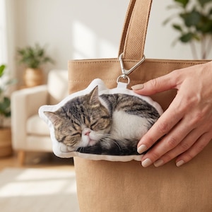 May include: A cat-shaped keyring showing a sleeping tabby cat with brown and white fur. The keyring is attached to a tan tote bag with a silver clasp. The cat's eyes are closed, and it appears to be resting comfortably.