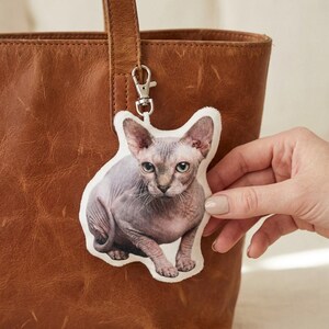 May include: A fabric keychain depicting a Sphynx cat with grey and pink skin, green eyes, and a white border. The keychain is attached to a silver clasp and is held near a brown leather tote bag.