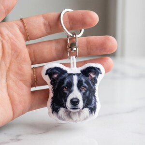 May include: A keychain with a black and white Border Collie dog portrait. The dog has brown eyes and a white muzzle. The keychain includes a silver metal ring and clasp.