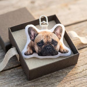May include: A French Bulldog plush keychain, presented in a brown gift box. The dog has brown and black fur, with a silver-colored clasp. The box is open, revealing the keychain resting on a cream-colored surface. The box is on a wooden surface.