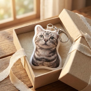 May include: A cat-shaped keyring with a silver clasp, presented in an open gift box tied with a cream ribbon. The keyring features a detailed image of a tabby cat with green eyes and a striped coat. The box is made of cardboard.