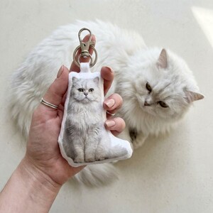 Op de afbeelding: Een wit en grijze kat sleutelhanger met een zilveren sluiting. De sleutelhanger heeft een gedetailleerde afbeelding van een pluizige kat. Een echte witte kat is op de achtergrond.