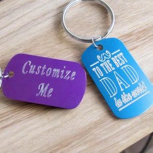 May include: Two metal keychains with engraved text. One keychain is purple and says "Customize Me". The other keychain is blue and says "To the Best Dad in the World".