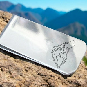 May include: Silver money clip with a wolf silhouette design. The clip is on a rock with a mountain background.