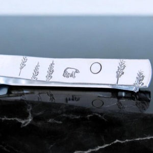 May include: A silver tie bar with an engraved design of a bear, a circle, and grass.