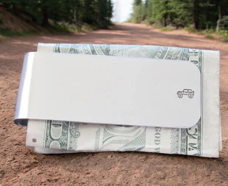 Peut inclure: Un porte-monnaie en argent avec une petite silhouette noire de Jeep sur le devant. Le porte-monnaie est en train de tenir un billet de dollar.