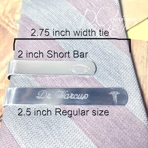 May include: Two silver tie bars on a striped tie. The top bar is 2 inches long and has a football design. The bottom bar is 2.5 inches long and has the text "Dr. Warcup" and a medical symbol.