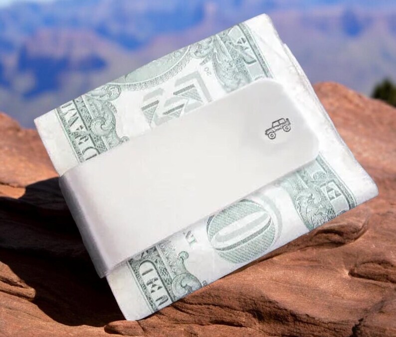 Peut inclure: Un clip &agrave; billets en m&eacute;tal blanc avec un petit dessin de voiture sur le devant. Le clip est en train de tenir une pile de billets de dollars am&eacute;ricains.