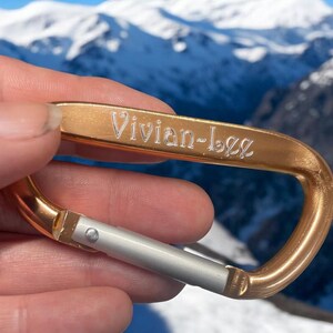 May include: A gold-colored carabiner with a silver latch. The carabiner is engraved with the name "Vivian-Lee".