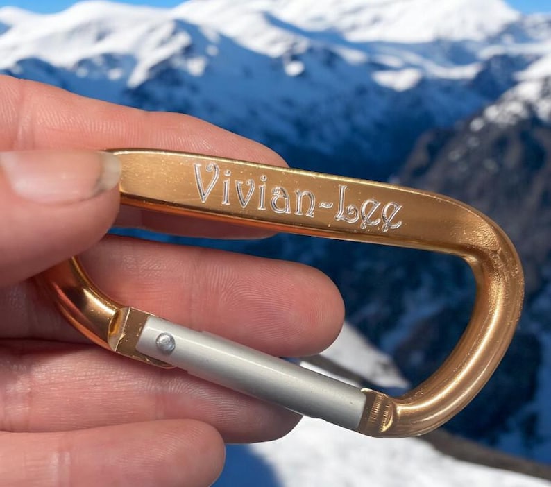 May include: A gold-colored carabiner with a silver latch. The carabiner is engraved with the name "Vivian-Lee".