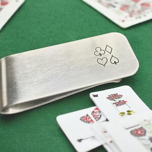 May include: A silver money clip with a design of four playing card suits: a diamond, a club, a heart, and a spade.
