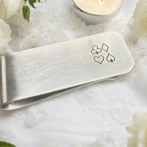 May include: Silver money clip with a brushed finish and an engraved design of four playing card suits: clubs, diamonds, hearts, and spades.