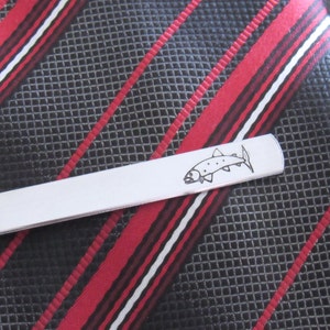 May include: Silver tie clip featuring a black and white outline of a fish. The tie clip is on a black and red striped tie.