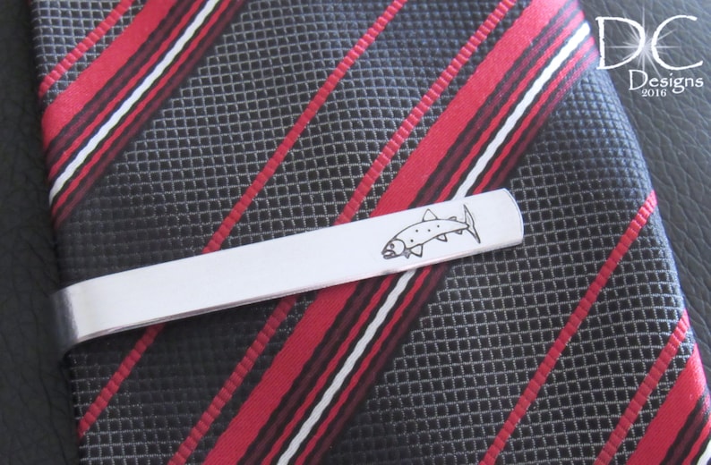 May include: Silver tie clip featuring a black and white outline of a fish. The tie clip is on a black and red striped tie.