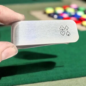 May include: A silver money clip with a design of four playing card suits: clubs, diamonds, hearts, and spades.