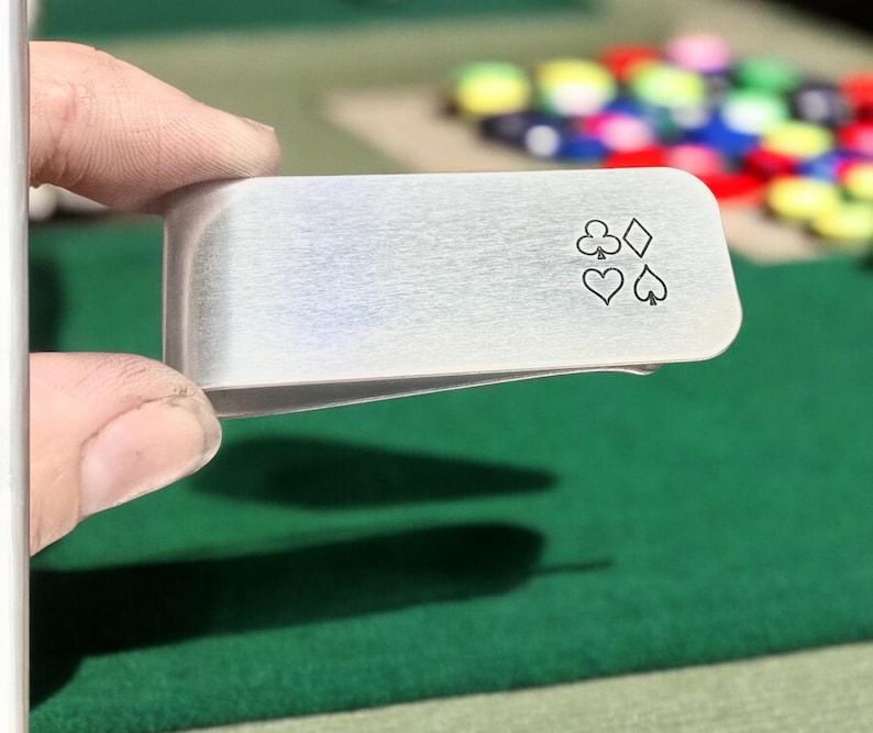 May include: A silver money clip with a design of four playing card suits: clubs, diamonds, hearts, and spades.