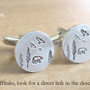 May include: A pair of silver cufflinks with a design of a bear, trees, and birds. The cufflinks are round and have a smooth surface.