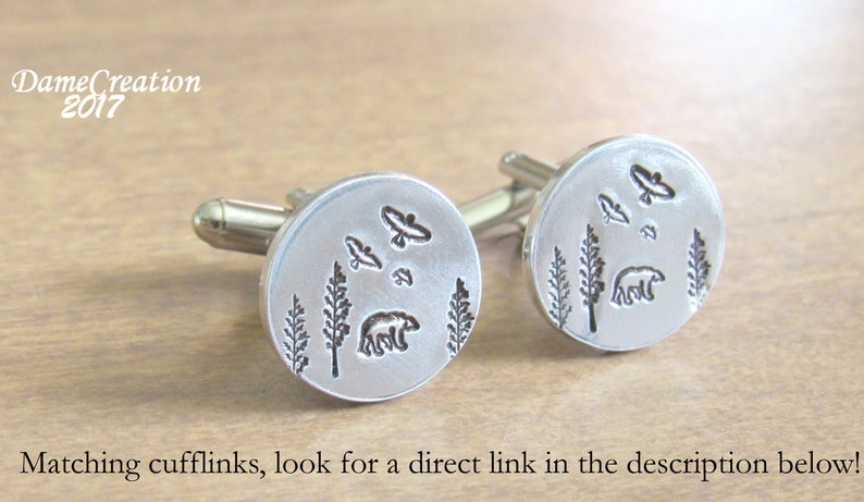 May include: A pair of silver cufflinks with a design of a bear, trees, and birds. The cufflinks are round and have a smooth surface.