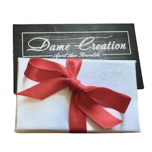 May include: A white gift box with a red ribbon and bow. The box has a label that says "Dame Creation - April Ann Kowalski".