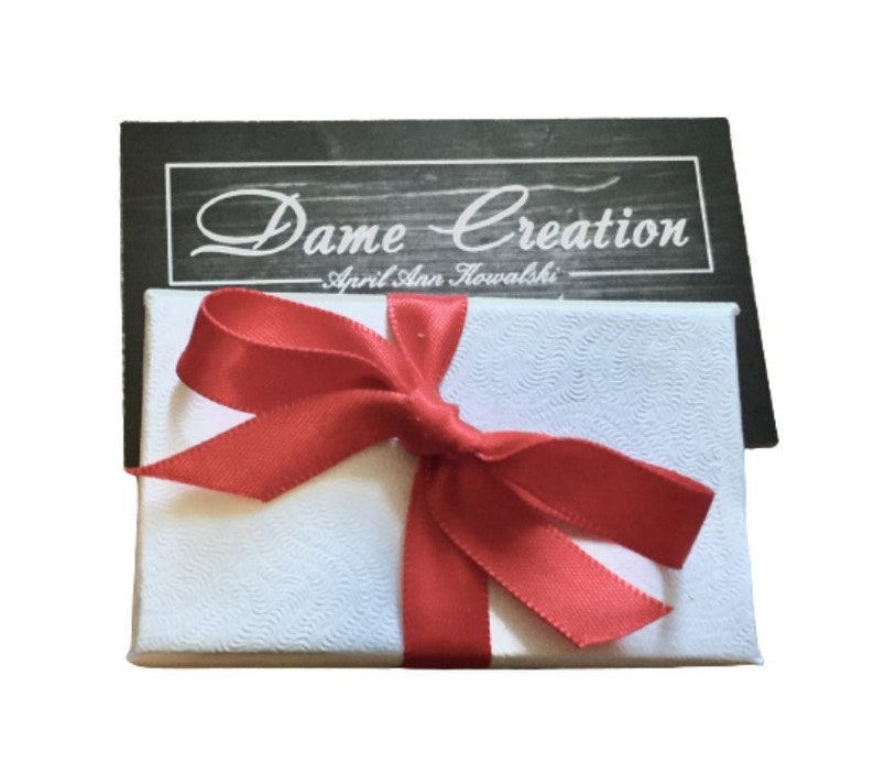 May include: A white gift box with a red ribbon and bow. The box has a label that says "Dame Creation - April Ann Kowalski".