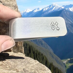 May include: A silver money clip with a design of four playing card suits: clubs, diamonds, hearts, and spades.