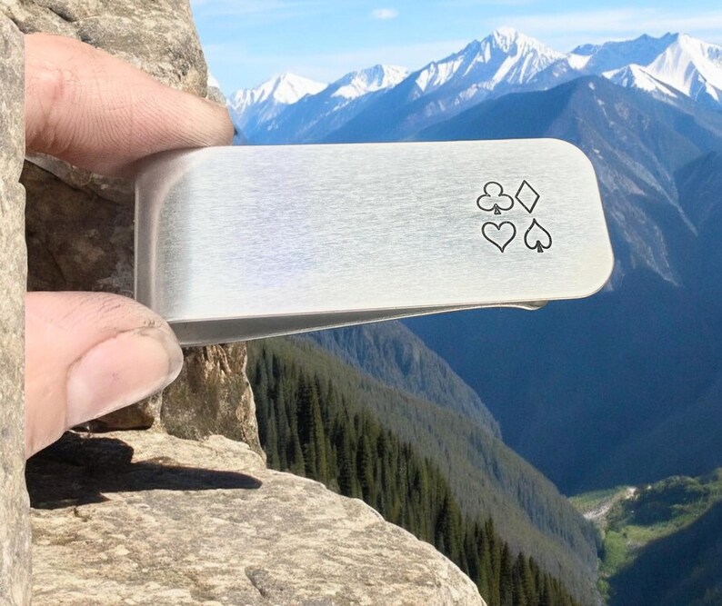 May include: A silver money clip with a design of four playing card suits: clubs, diamonds, hearts, and spades.