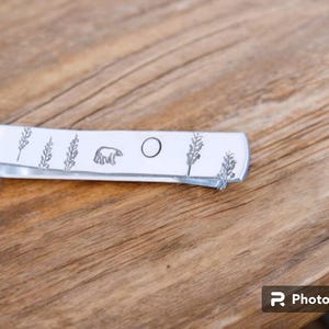 May include: A silver metal tie bar with a design of a bear, a circle, and pine trees.