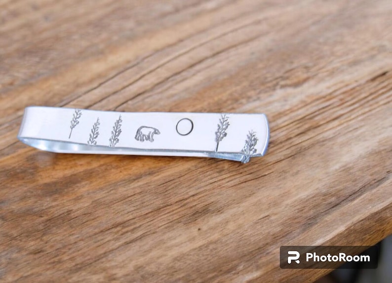 May include: A silver metal tie bar with a design of a bear, a circle, and pine trees.