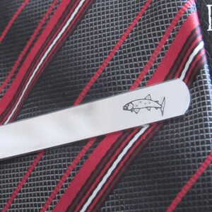 May include: Silver tie clip featuring a black and white outline of a fish. The tie clip is on a red and black striped tie.
