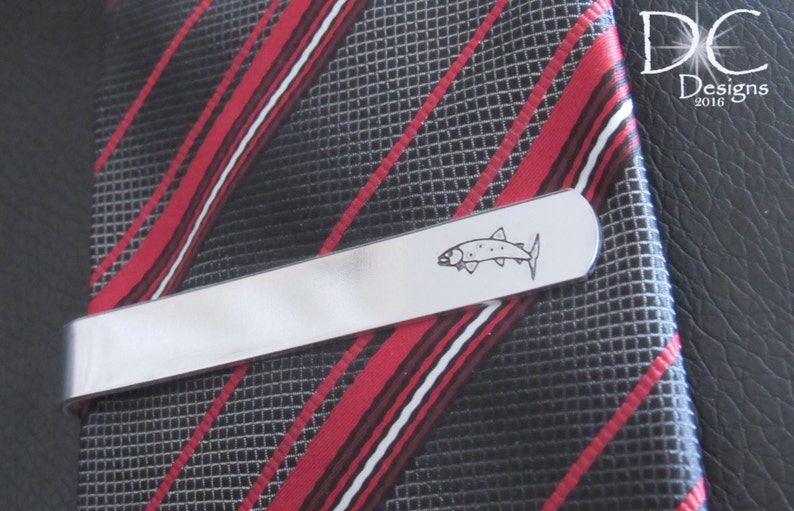 May include: Silver tie clip featuring a black and white outline of a fish. The tie clip is on a red and black striped tie.