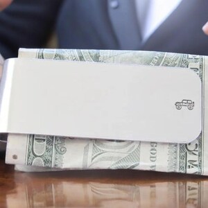 Peut inclure: Un porte-monnaie en argent avec un petit dessin de voiture sur le devant. Le porte-monnaie contient une pile de billets de dollars am&eacute;ricains.