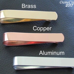 K&ouml;nnte beinhalten: Drei Geldclips aus Metall in verschiedenen Ausf&uuml;hrungen: Messing, Kupfer und Aluminium. Die Clips sind vertikal auf einer schwarzen Lederoberfl&auml;che angeordnet.