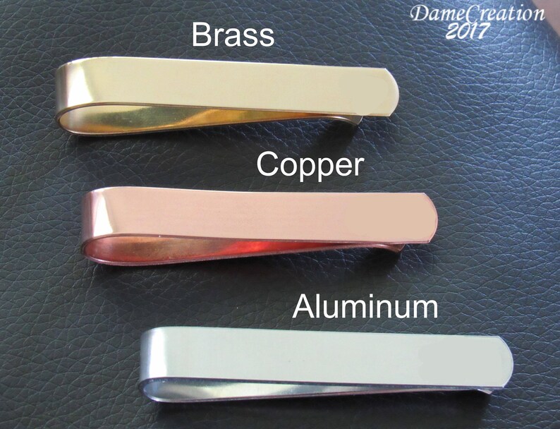 K&ouml;nnte beinhalten: Drei Geldclips aus Metall in verschiedenen Ausf&uuml;hrungen: Messing, Kupfer und Aluminium. Die Clips sind vertikal auf einer schwarzen Lederoberfl&auml;che angeordnet.