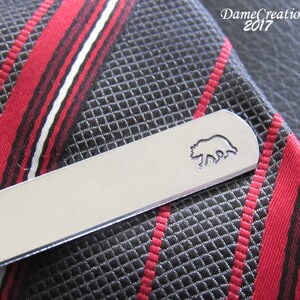 May include: Silver tie bar with a bear silhouette design. The tie bar is lying on a black and red striped tie.