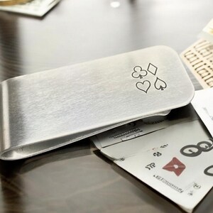 May include: A silver money clip with a design of four playing card suits: clubs, diamonds, hearts, and spades.