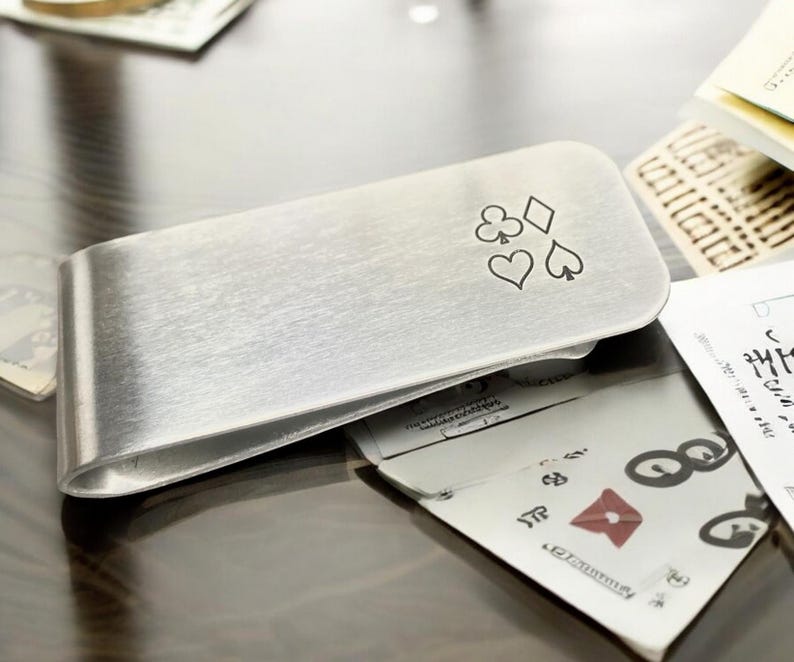May include: A silver money clip with a design of four playing card suits: clubs, diamonds, hearts, and spades.