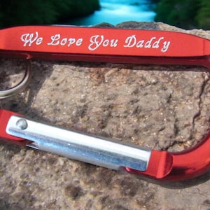 May include: Red metal carabiner with a silver spring-loaded gate. The carabiner is engraved with the words "We Love You Daddy".