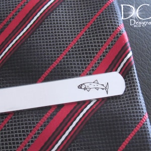 May include: Silver tie clip featuring a fish design. The tie clip is on a black and red striped tie.