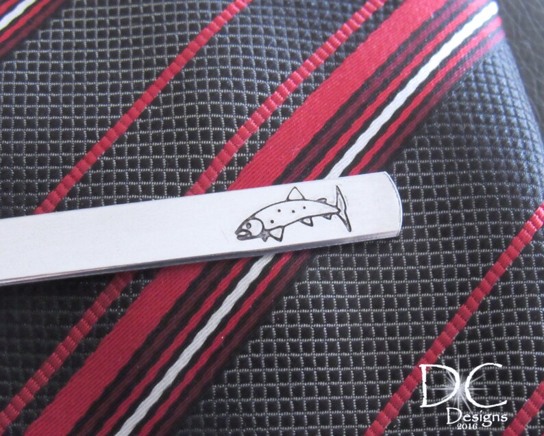 May include: Silver tie bar with an engraved fish design. The tie bar is on a black and red striped fabric.