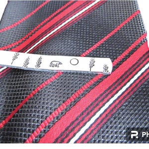 May include: Silver tie clip with a bear, a circle, and two sprigs of wheat engraved on it. The tie clip is on a red and black striped tie.
